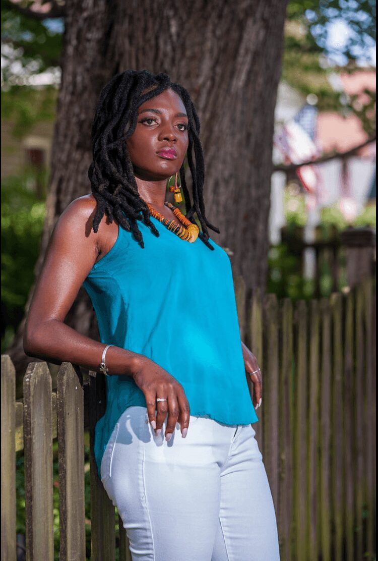Woman with dreadlocks wearing a blue top and white pants outdoors, leaning on a wooden fence near a tree.