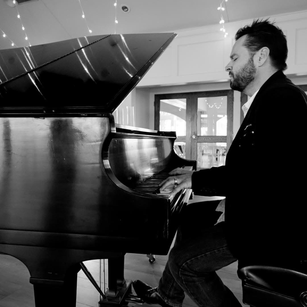 Man playing piano indoors, side view, black and white photo, focused expression, window in background.