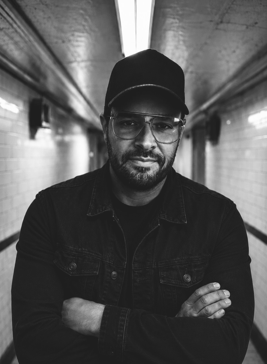 Man with glasses, beard, wearing a cap and jacket, standing with arms crossed in a tunnel, black and white photo.