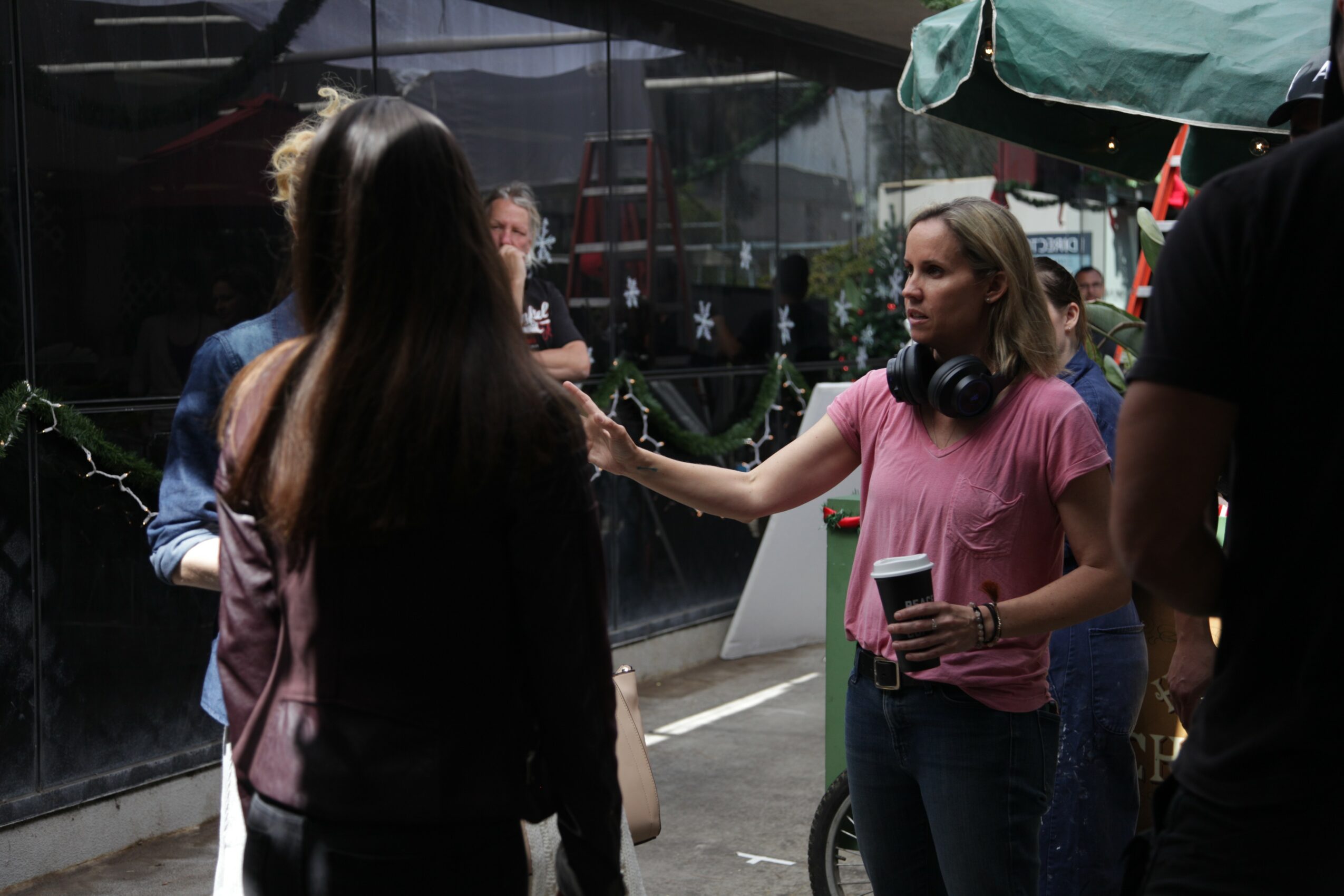 Group of people talking outdoors, woman with headphones holding a cup, others facing her, glass building in background.