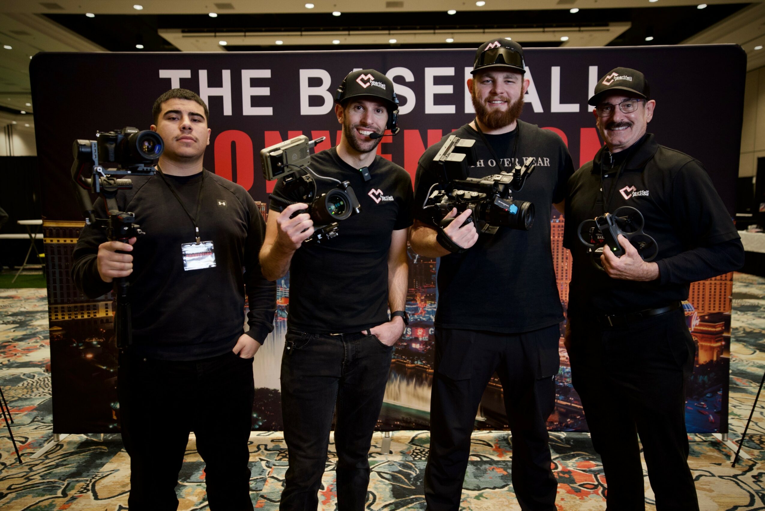 Four men holding cameras and equipment in front of a backdrop with text, indoors. All wear black clothing and caps.