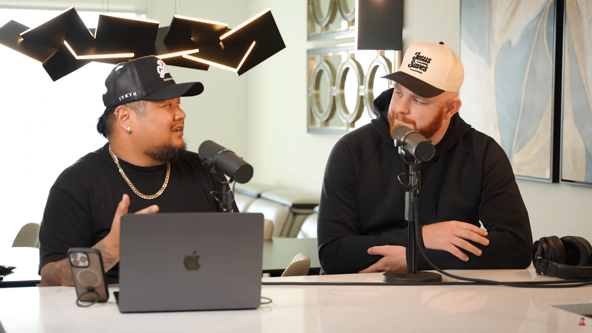 Two men sit at a table with microphones, a laptop, and headphones, engaged in conversation in a modern room.