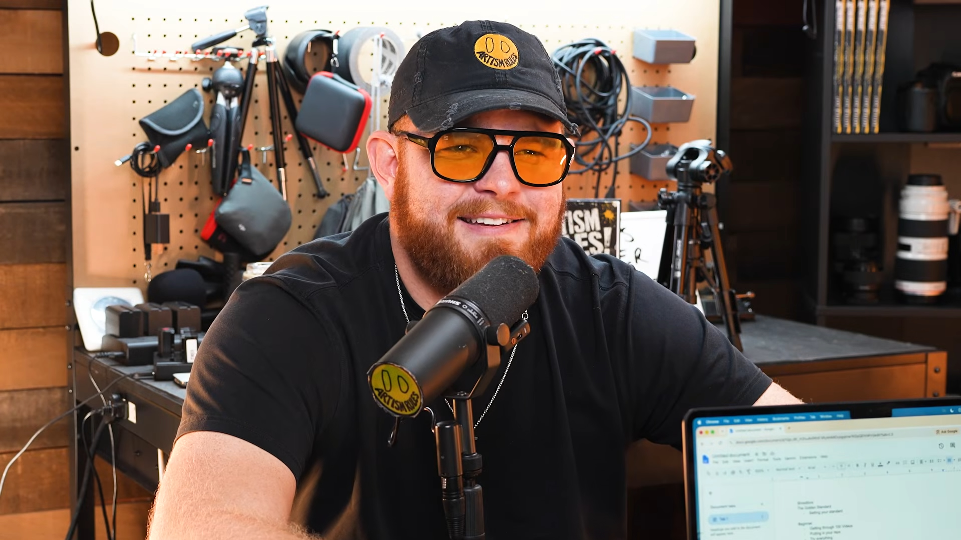 Smiling man with glasses and a cap speaking into a microphone in a recording studio, surrounded by equipment and shelves.