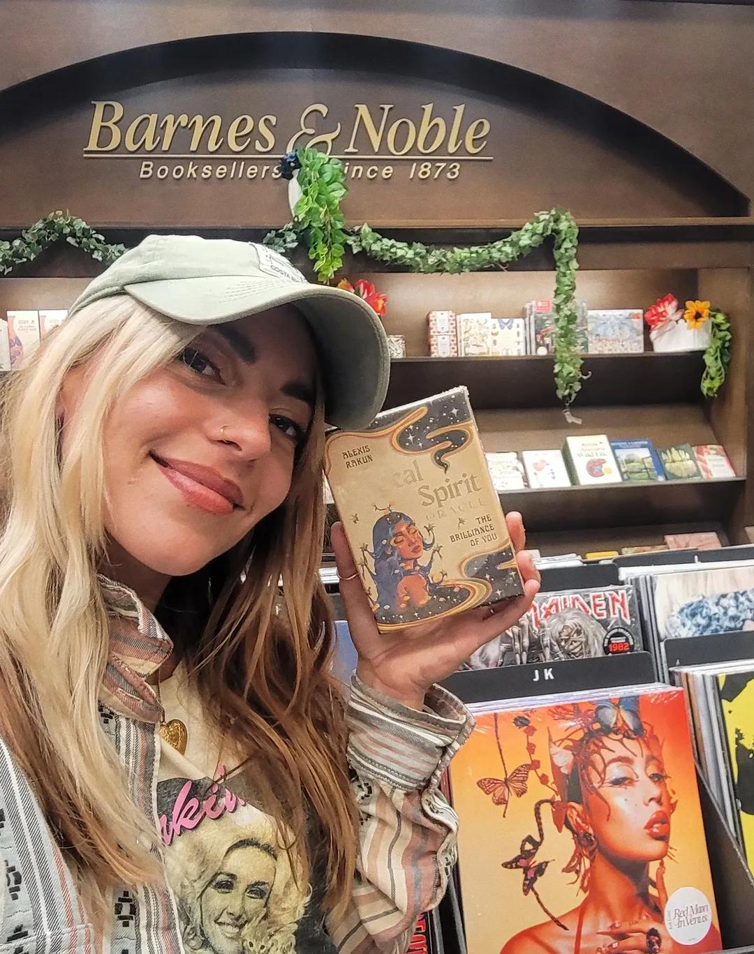 Woman with long blonde hair and a light-colored hat smiling inside Barnes & Noble bookstore, holding a book titled 'Spirit'.