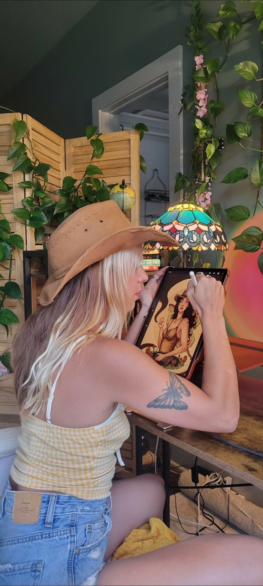 Person with long blonde hair, wearing a beige hat and yellow top, looking at a tablet in a decorated room.