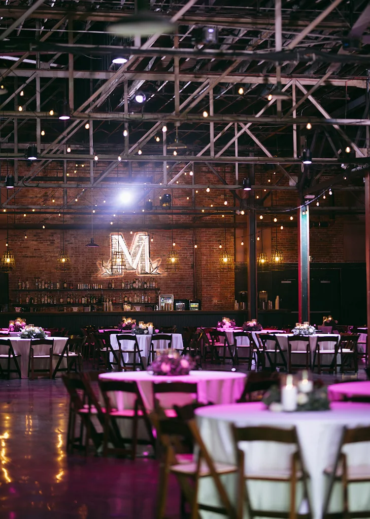 Event space with round tables, chairs, and a lit brick wall with a large letter 'M' in the background.