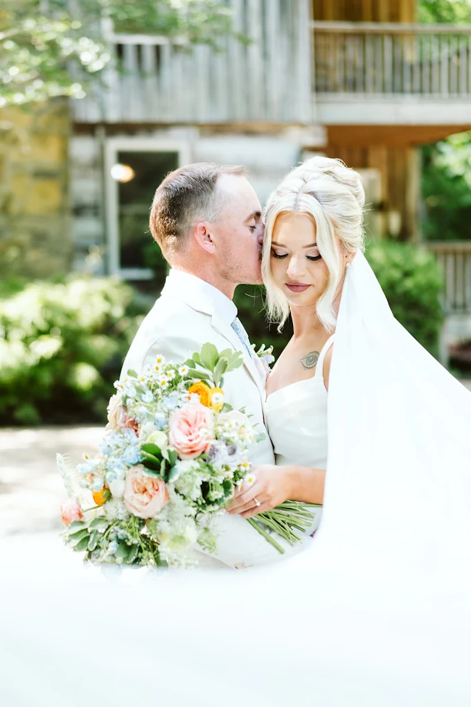 A bride and groom embrace outdoors, with the groom kissing the bride's forehead, holding a bouquet of flowers.