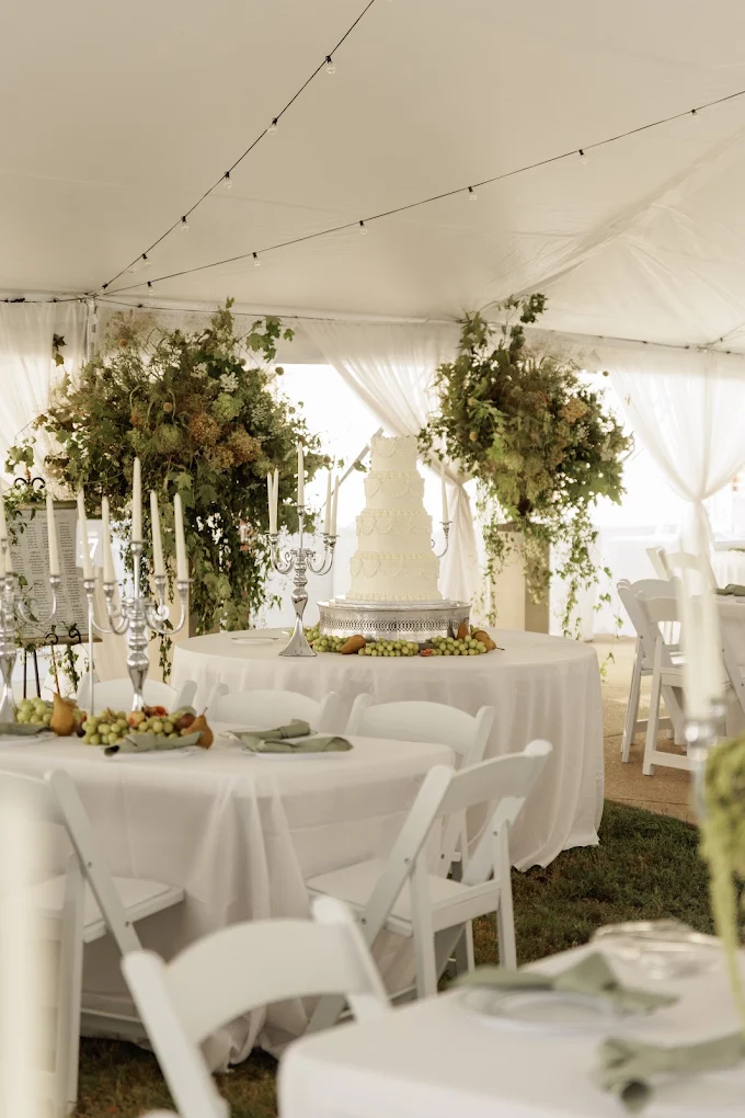 Elegant event setup with round tables, white chairs, floral centerpieces, and draped tent interior.