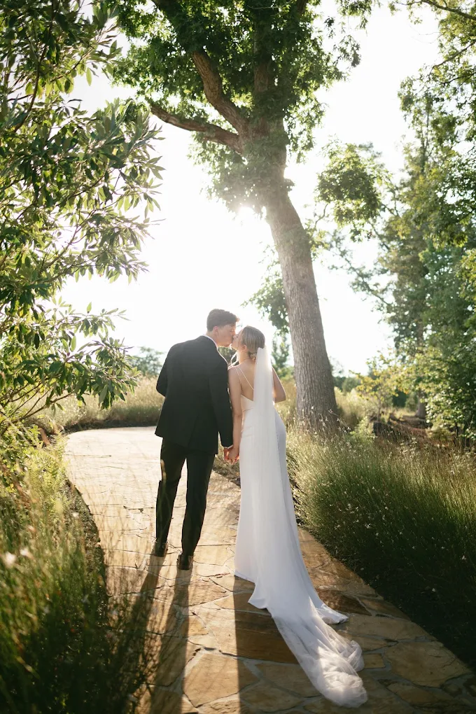 A bride and groom stand on a stone path surrounded by trees and greenery, sunlight shining through the leaves.