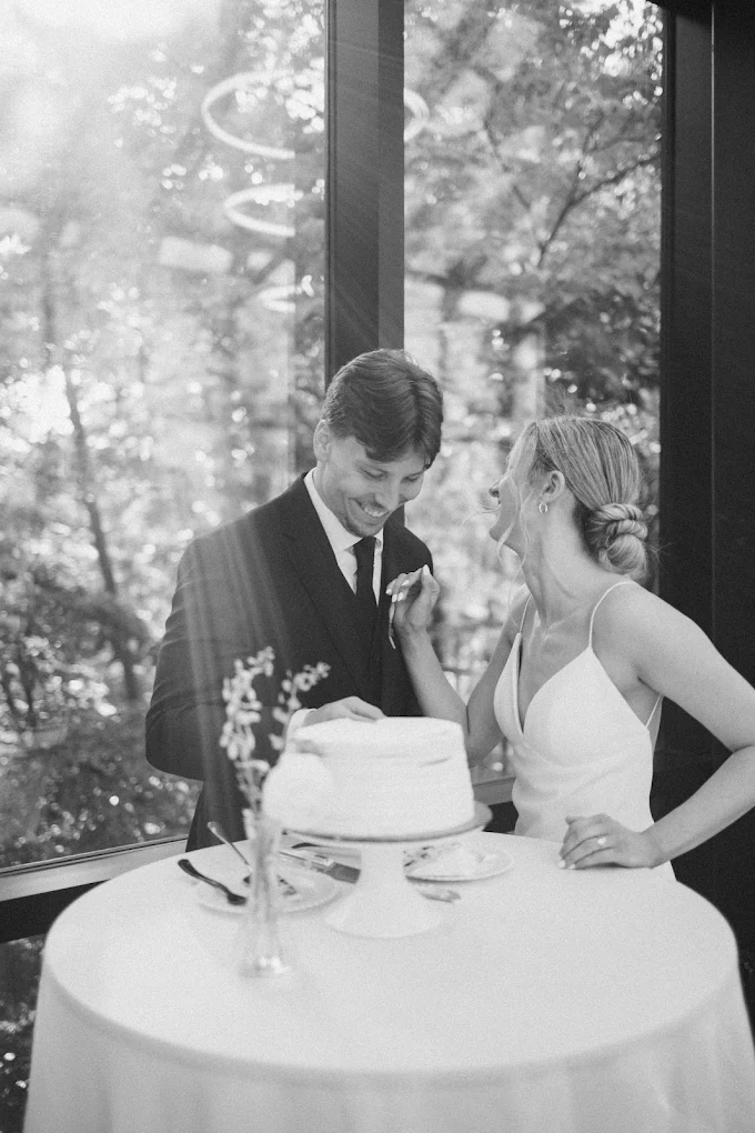 Two people, a man and a woman, stand near a table with a cake inside a room with large windows and trees outside.