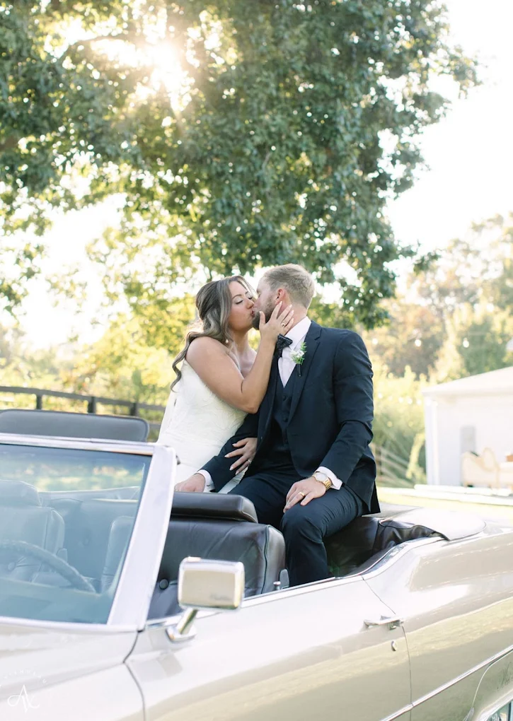 Couple in wedding attire sharing a kiss on a convertible car outdoors with trees and sunlight.