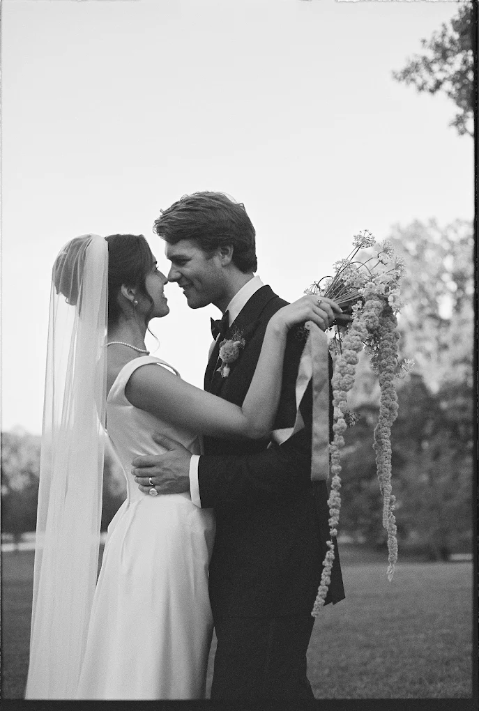 Bride and groom face each other outdoors, holding a bouquet, smiling, with trees in the background.
