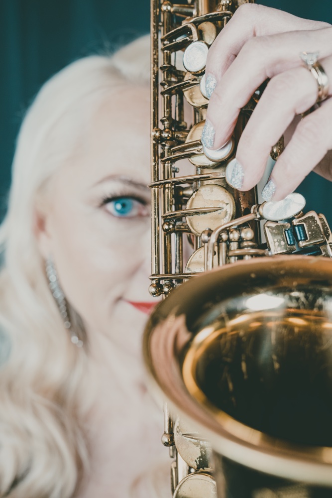 Close-up of a woman with blonde hair and blue eyes holding a saxophone, partially covering her face.