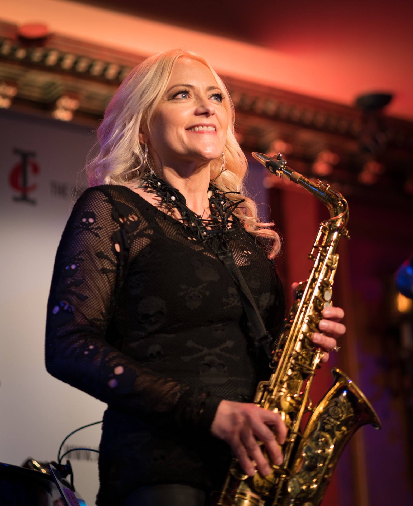Woman with blonde hair holding a saxophone, smiling on stage with warm lighting.