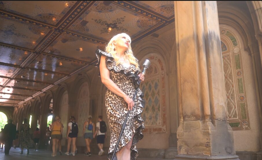 Person with long blonde hair in a leopard print dress holding a microphone, standing in ornate architectural setting.