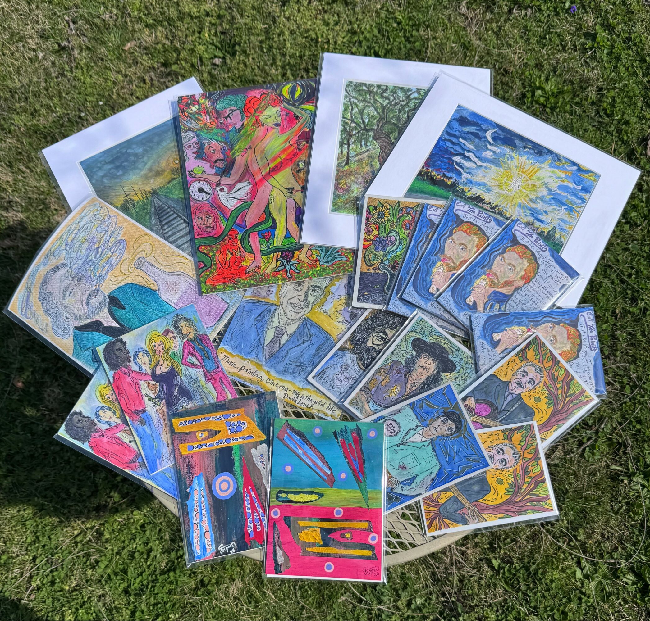 Collection of various colorful art prints and paintings spread on grass, including landscapes, portraits, and abstract designs.