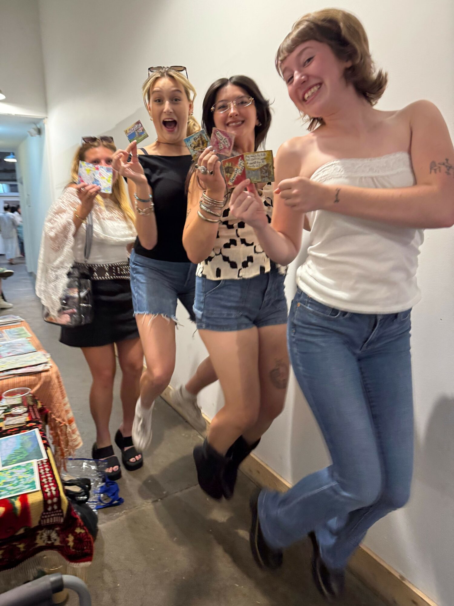 Four women taking a mirror selfie in a room, smiling and posing, with a table and colorful items in the foreground.