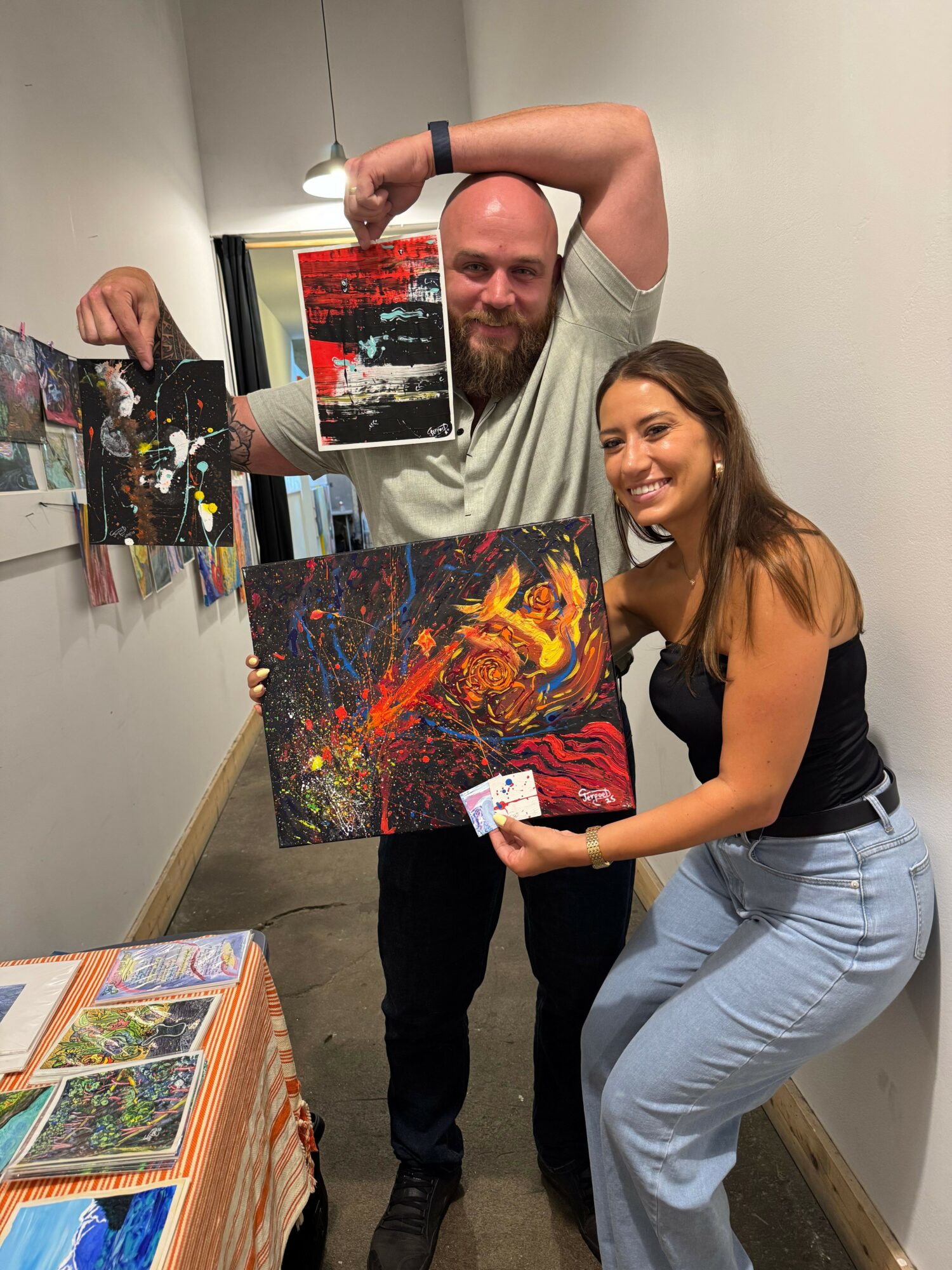 Two people, one holding a colorful abstract painting, stand indoors with a table of artwork in the foreground.