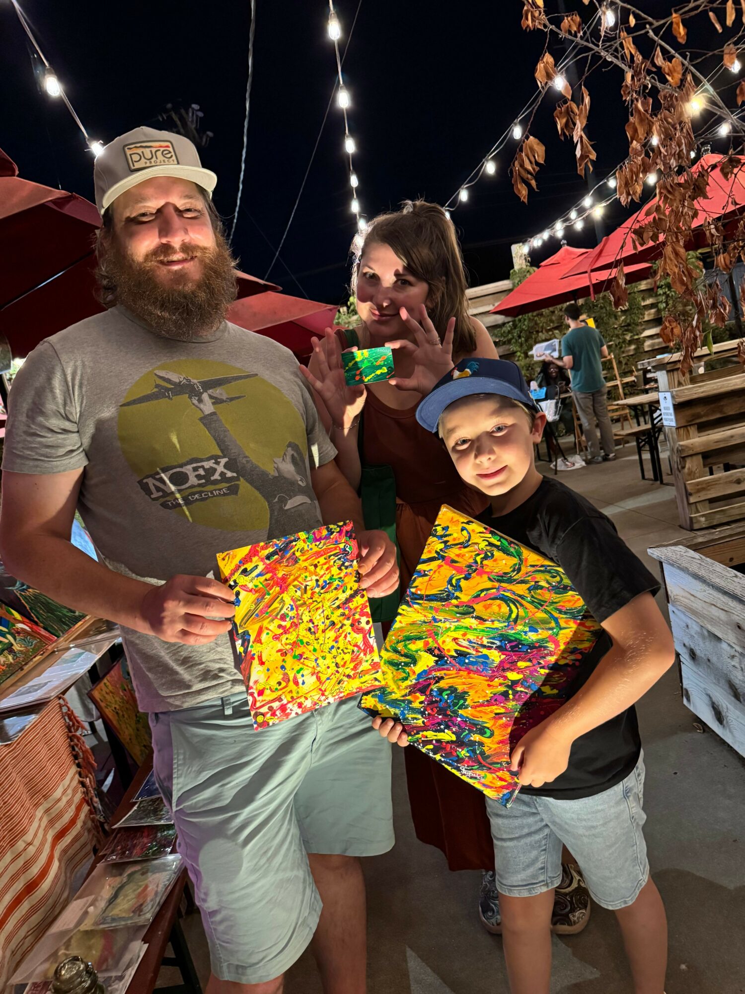 Two men holding colorful abstract paintings outdoors at night, with string lights and trees in the background.