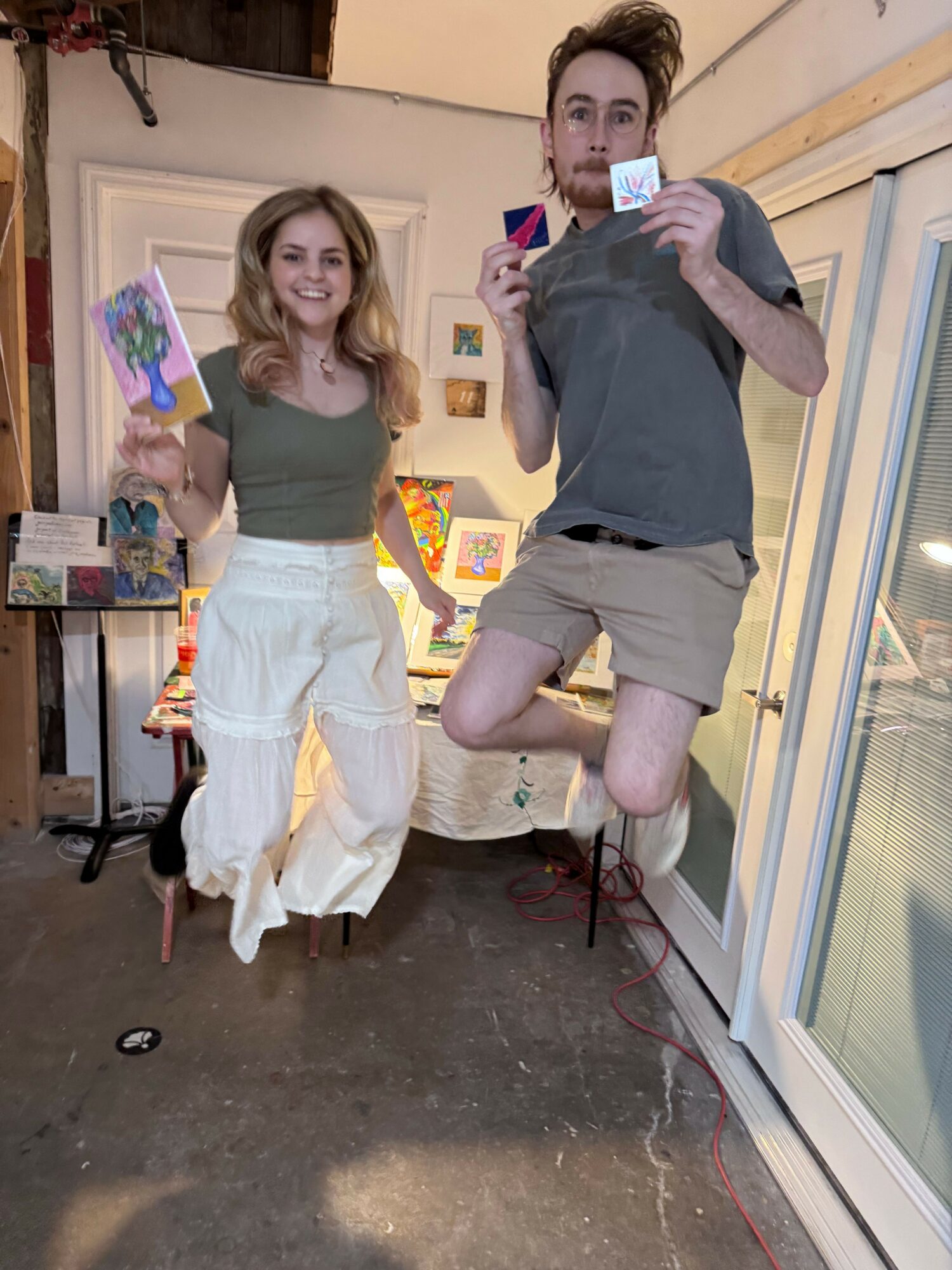 Two women standing indoors, one holding a phone, with colorful artwork and a window behind them.
