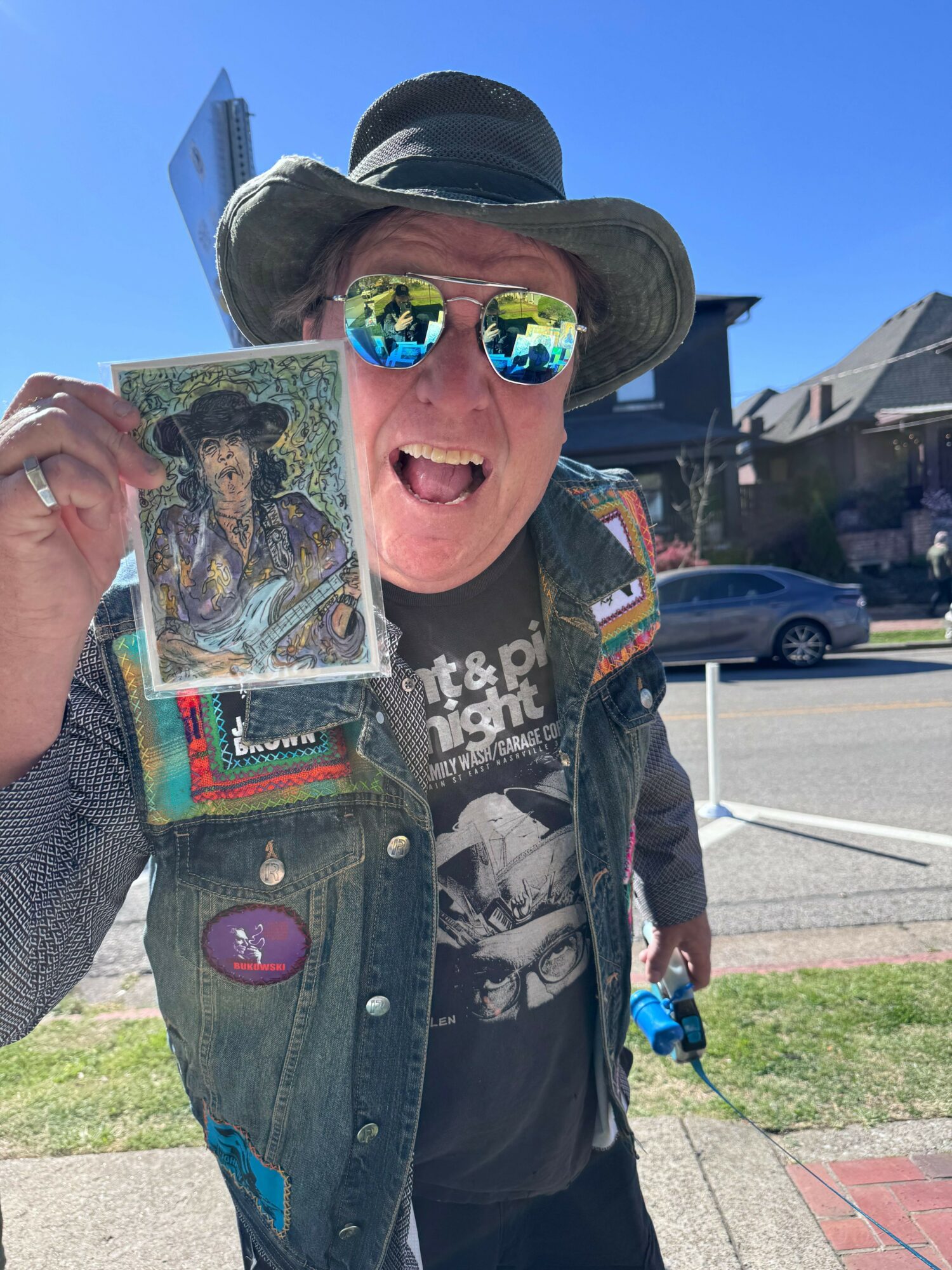 Person wearing sunglasses and a hat, holding a religious icon, smiling outdoors on a sunny day.