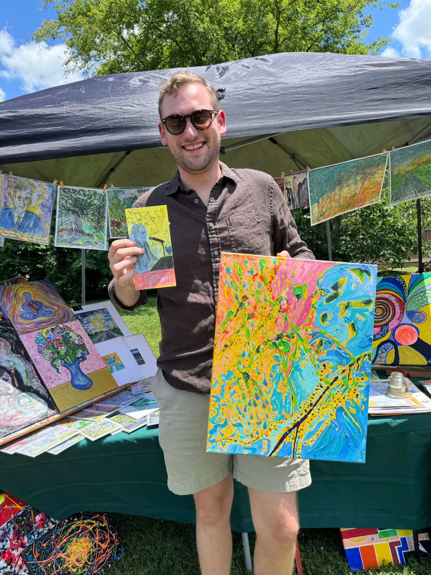 Person with sunglasses holding a colorful painting at outdoor art display, smiling, with trees and artwork in background.
