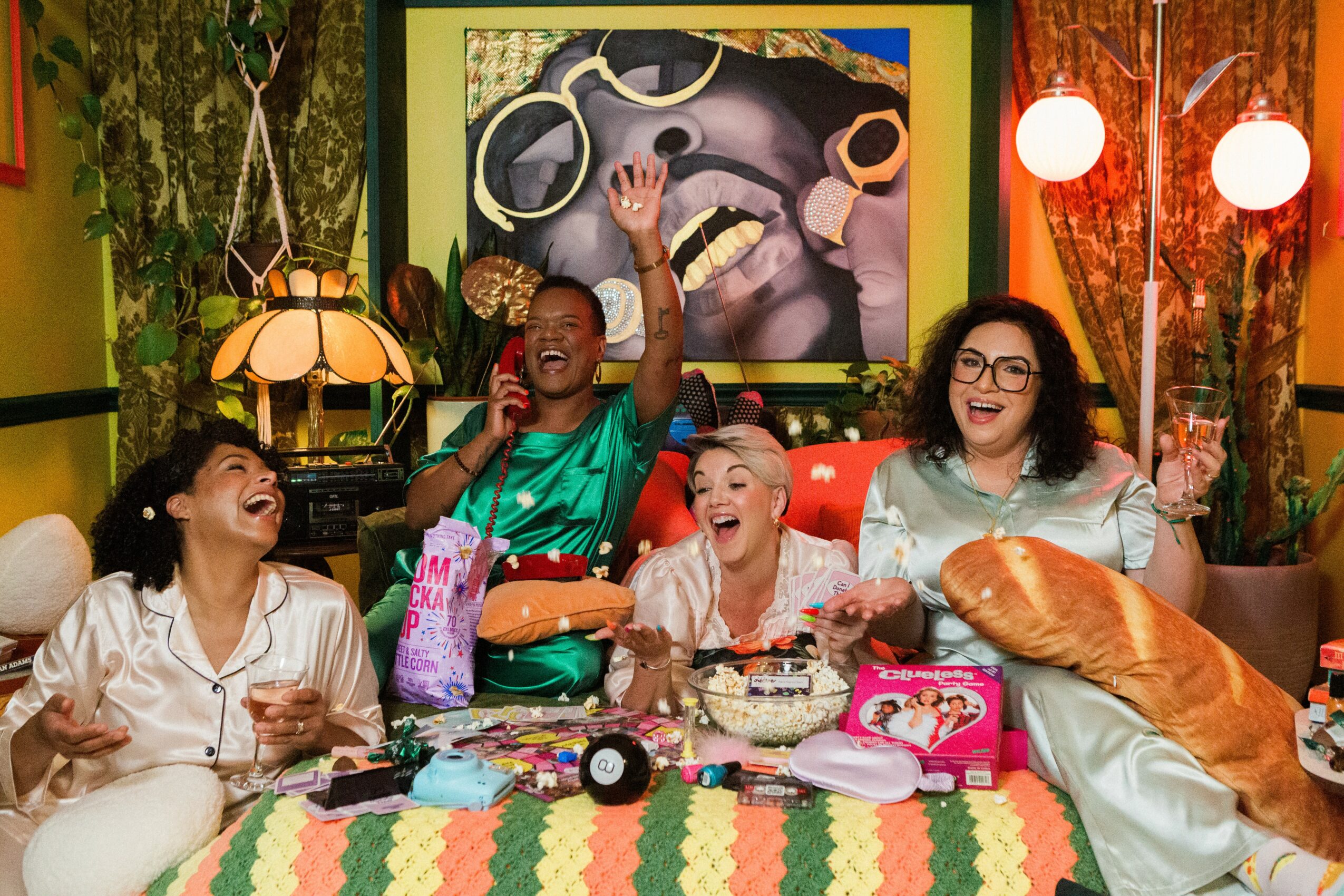 Group of five women celebrating, laughing, and raising glasses in a colorful, decorated room with artwork and lamps.