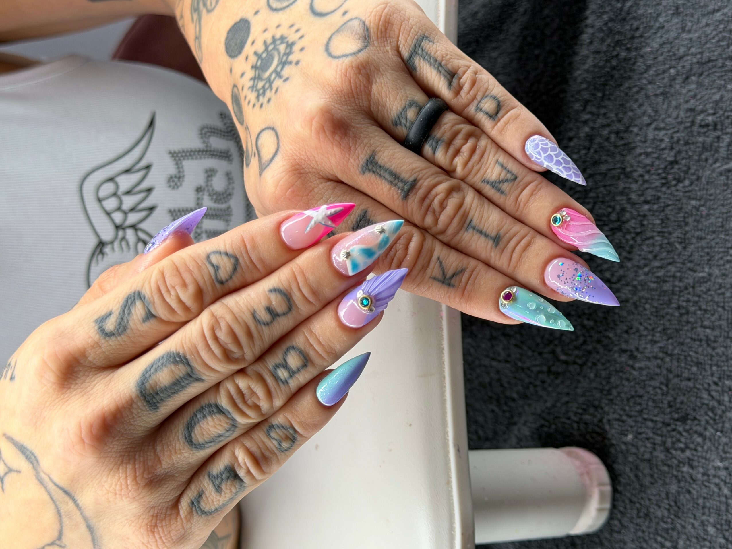 Close-up of hands with colorful, decorated nails and tattoos, resting on a surface with a tattooed arm visible.
