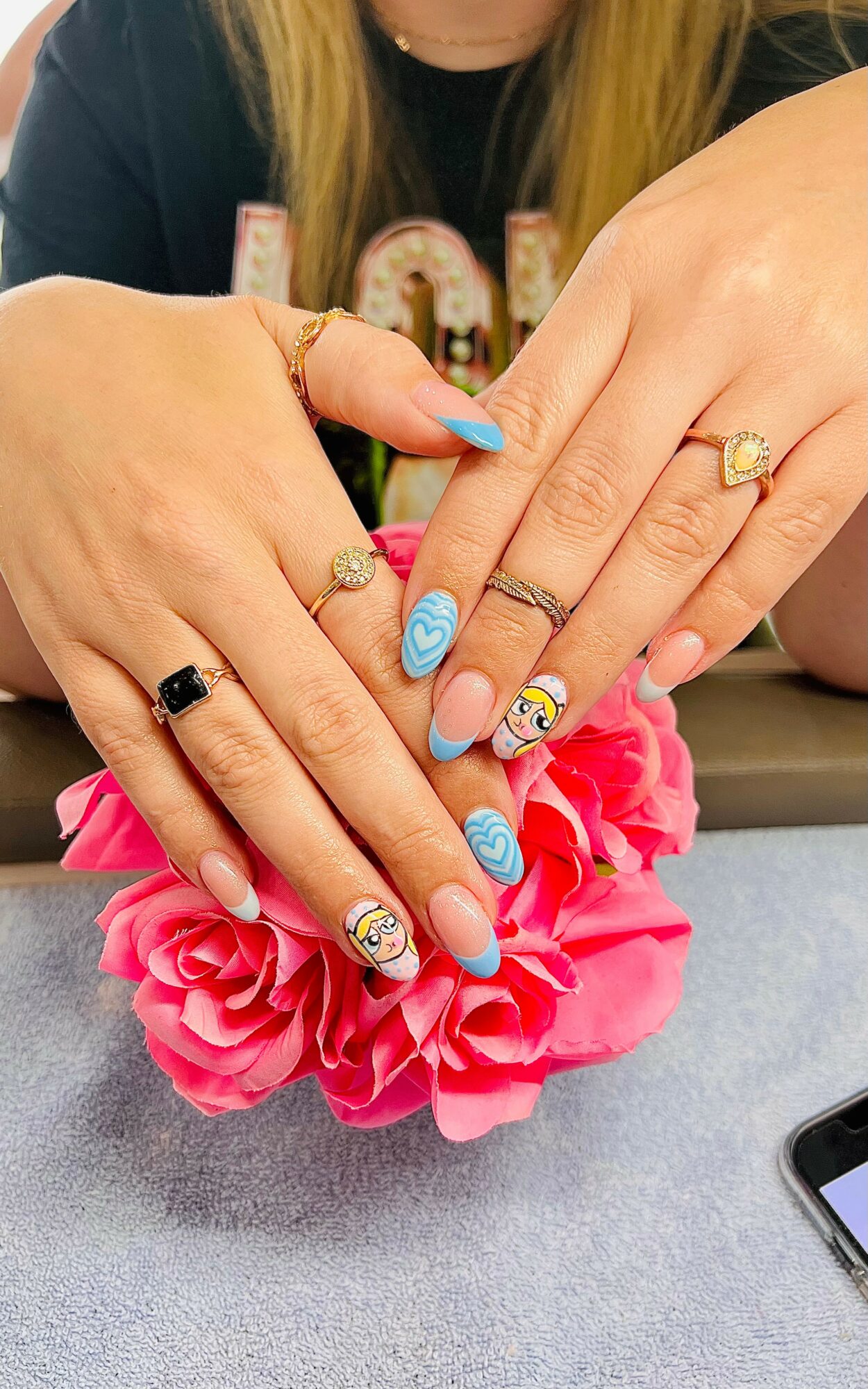 Two hands with decorated nails resting on pink flower, rings on fingers, background of person with long hair and dark shirt.