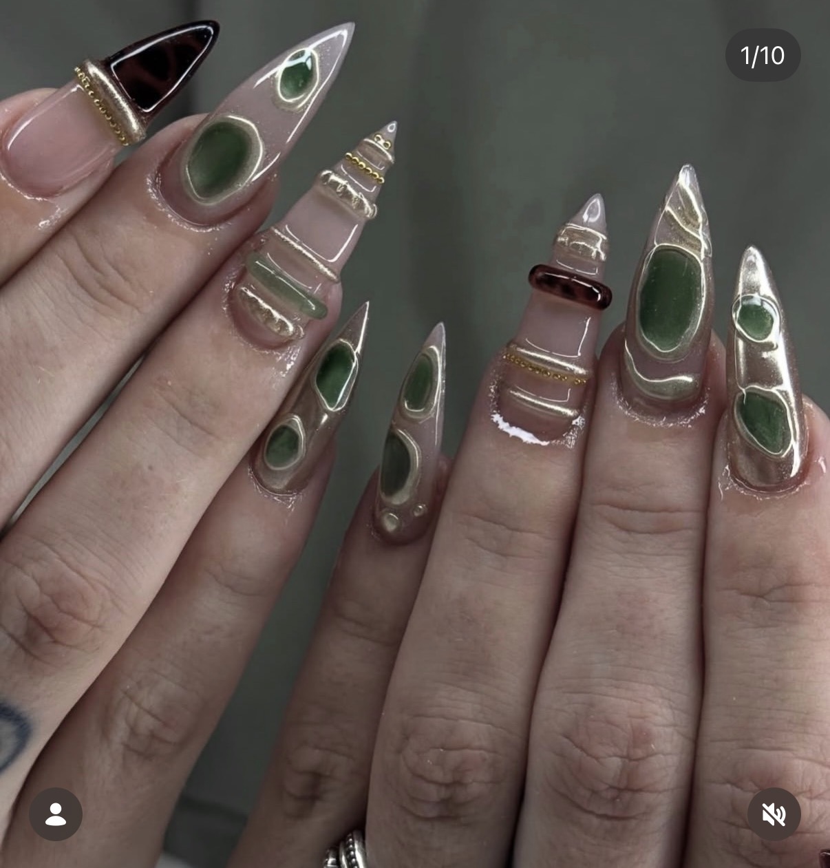 Close-up of long, pointed, metallic nails with green and black accents, held by fingers with rings, against a plain background.