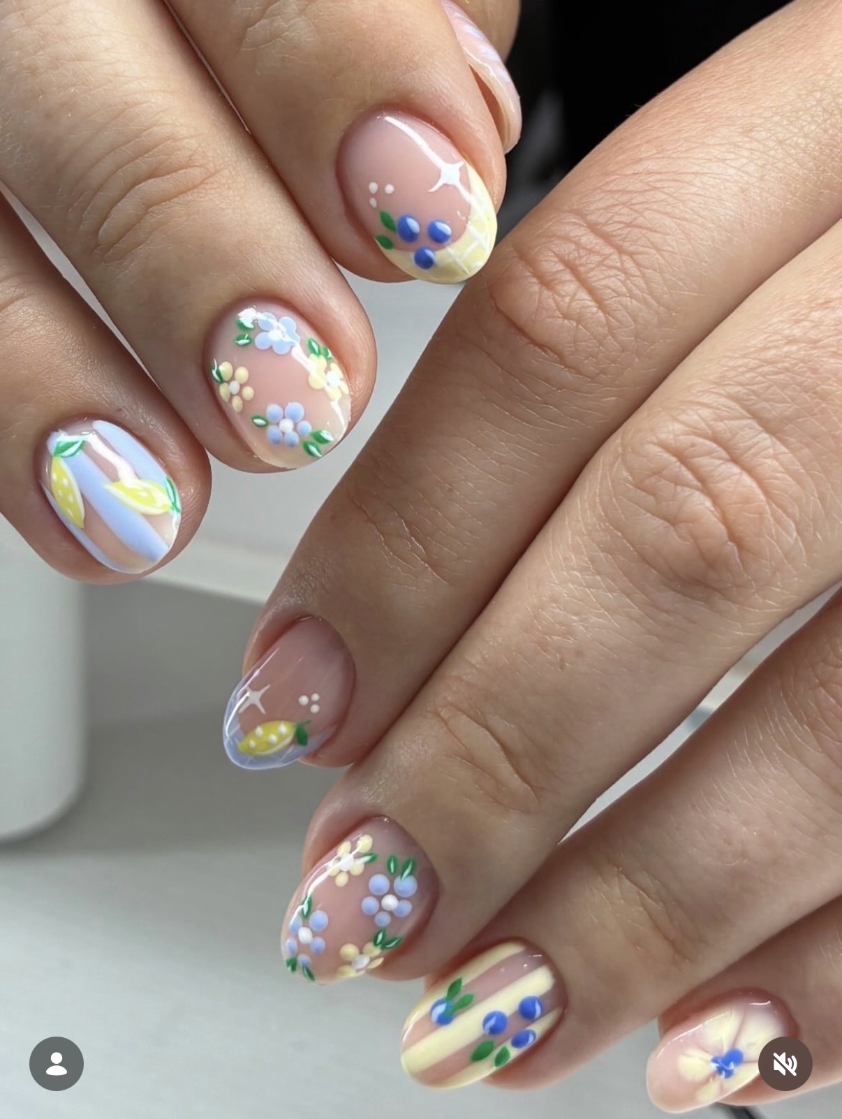 Close-up of hands with decorated nails featuring floral, rainbow, and star designs on a clear base, with a blurred background.