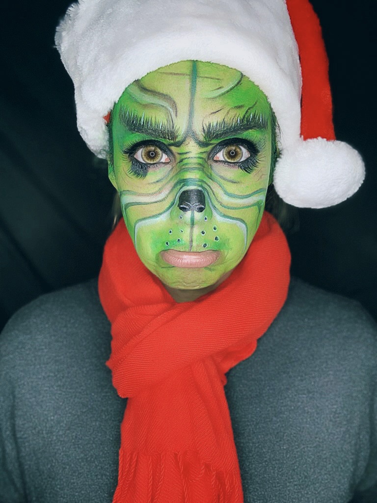 Person with green face paint resembling a cat, wearing a Santa hat and red scarf, looking directly at the camera.