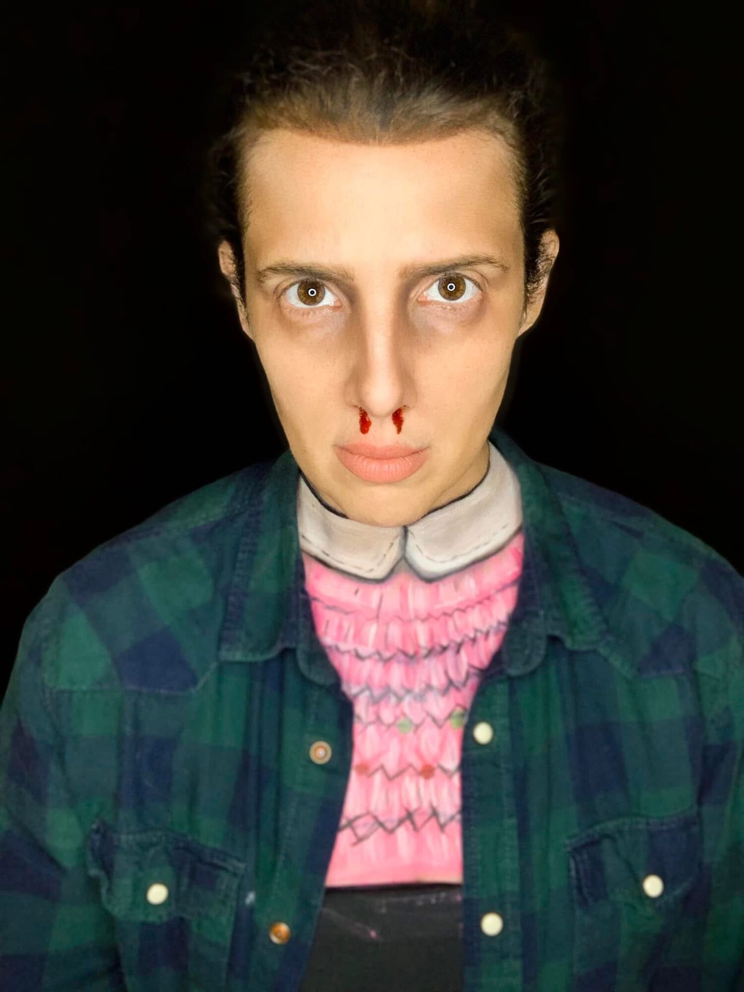 Person with nosebleed, wearing a pink sweater and green plaid shirt, against a black background.