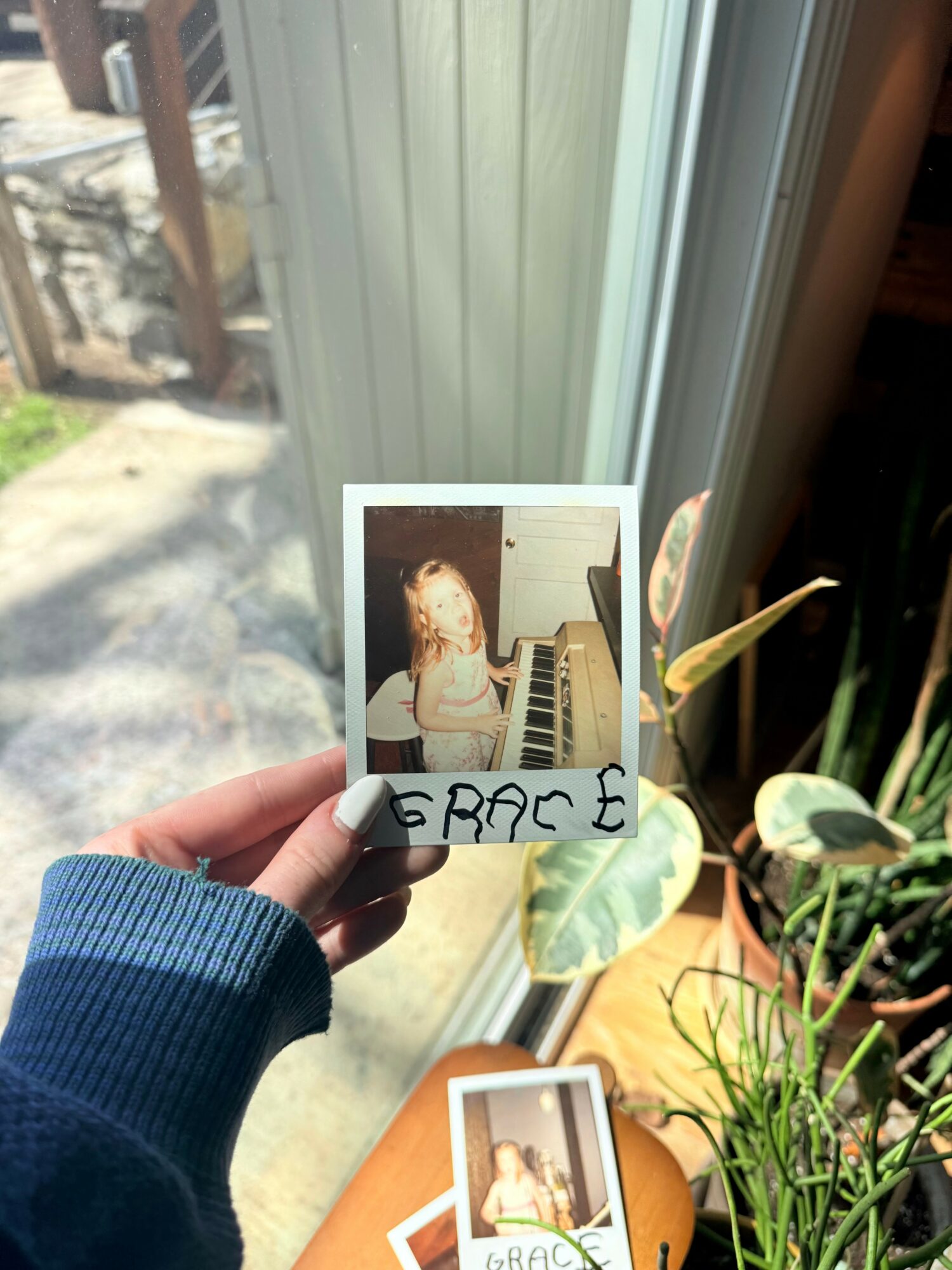Person holding a photo of a girl with long hair sitting at a piano, with handwritten name 'GRACE' on the bottom, near a window with plants.