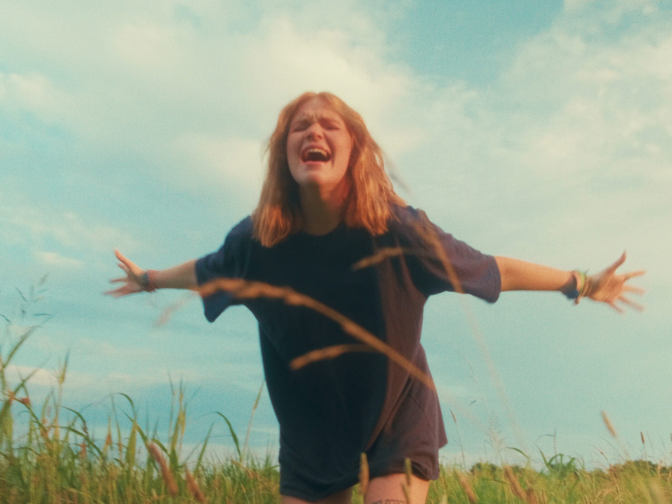 Woman with arms outstretched, smiling in a grassy field under a partly cloudy sky.