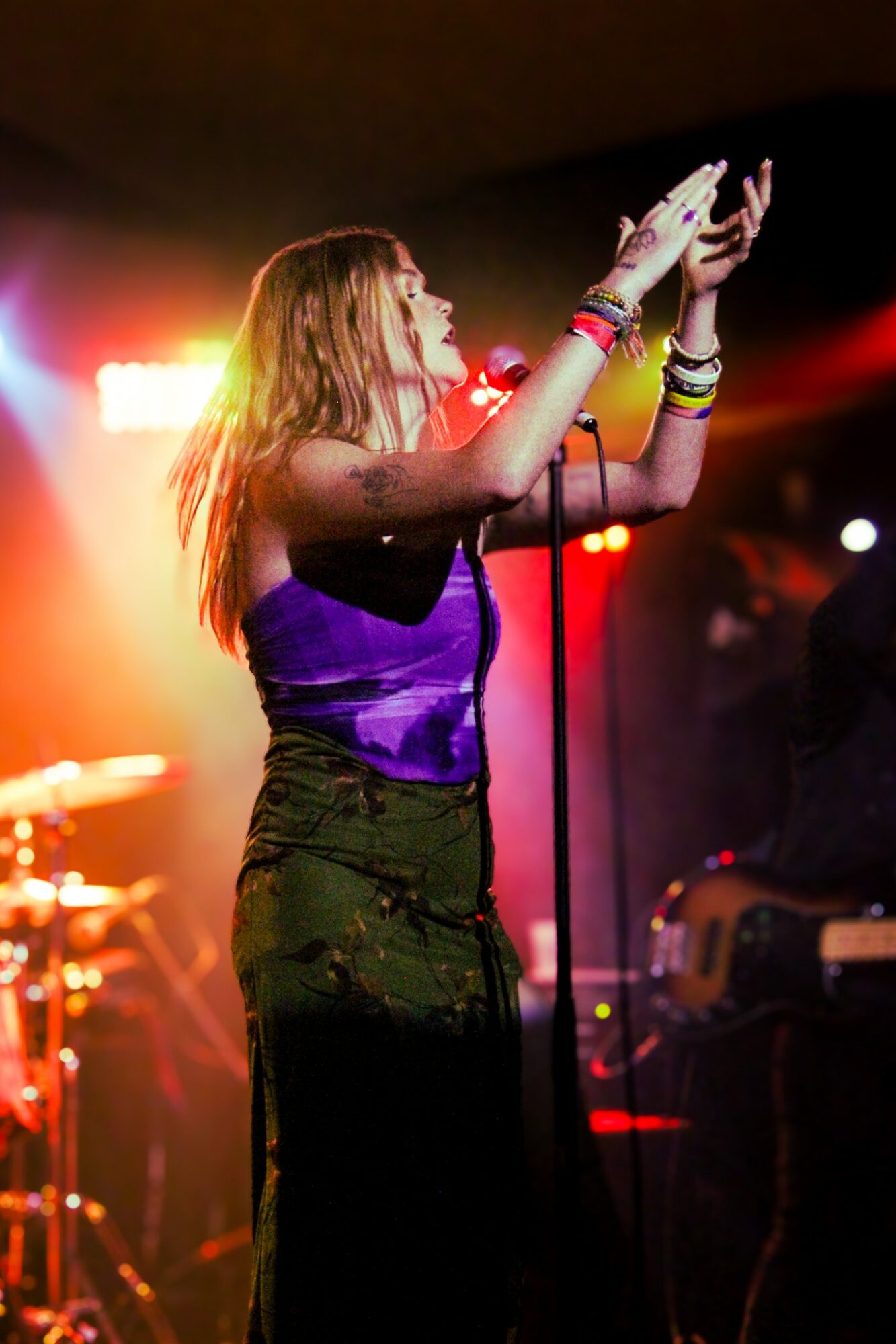 Woman singing into a microphone on stage with colorful lights, wearing a purple top and accessories.