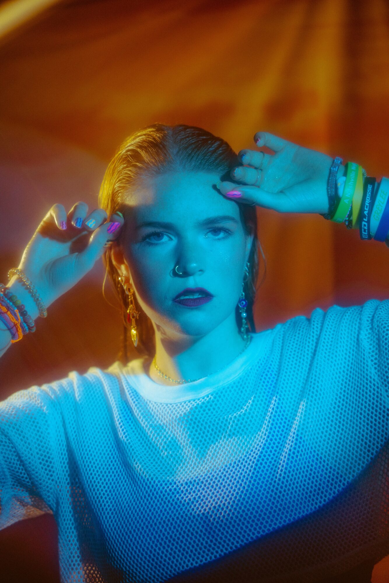 Young woman with braided hair and colorful bracelets, posing with hands near her face, colorful lighting background.