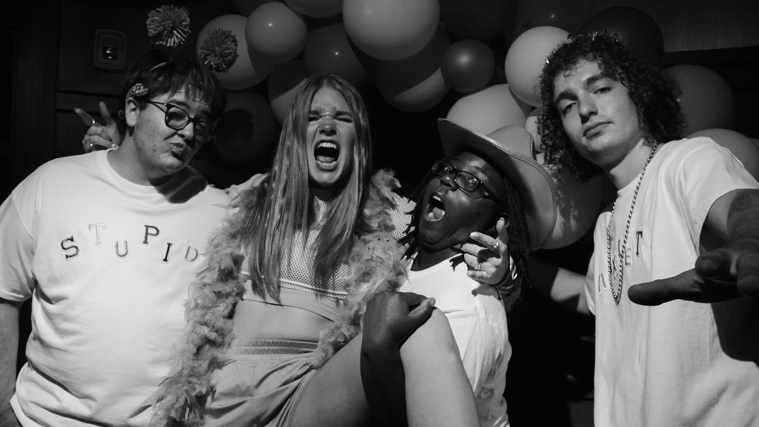 Four people posing together, some making expressive gestures, with balloons in the background.