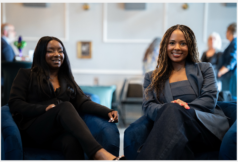 Two women sitting on chairs, smiling, in a bright indoor setting with other people in the background.