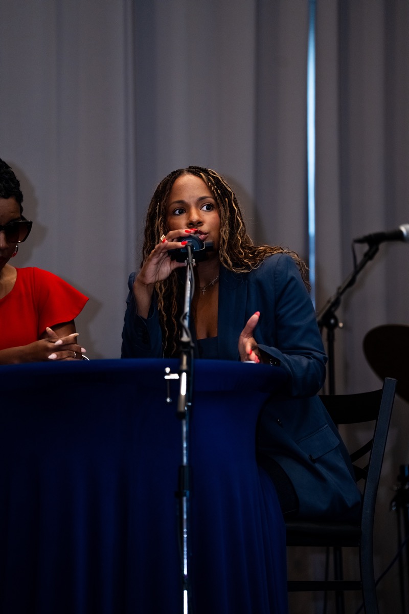 Woman with long curly hair speaking into a microphone at a panel, with a person in red beside her, in a room with curtains.