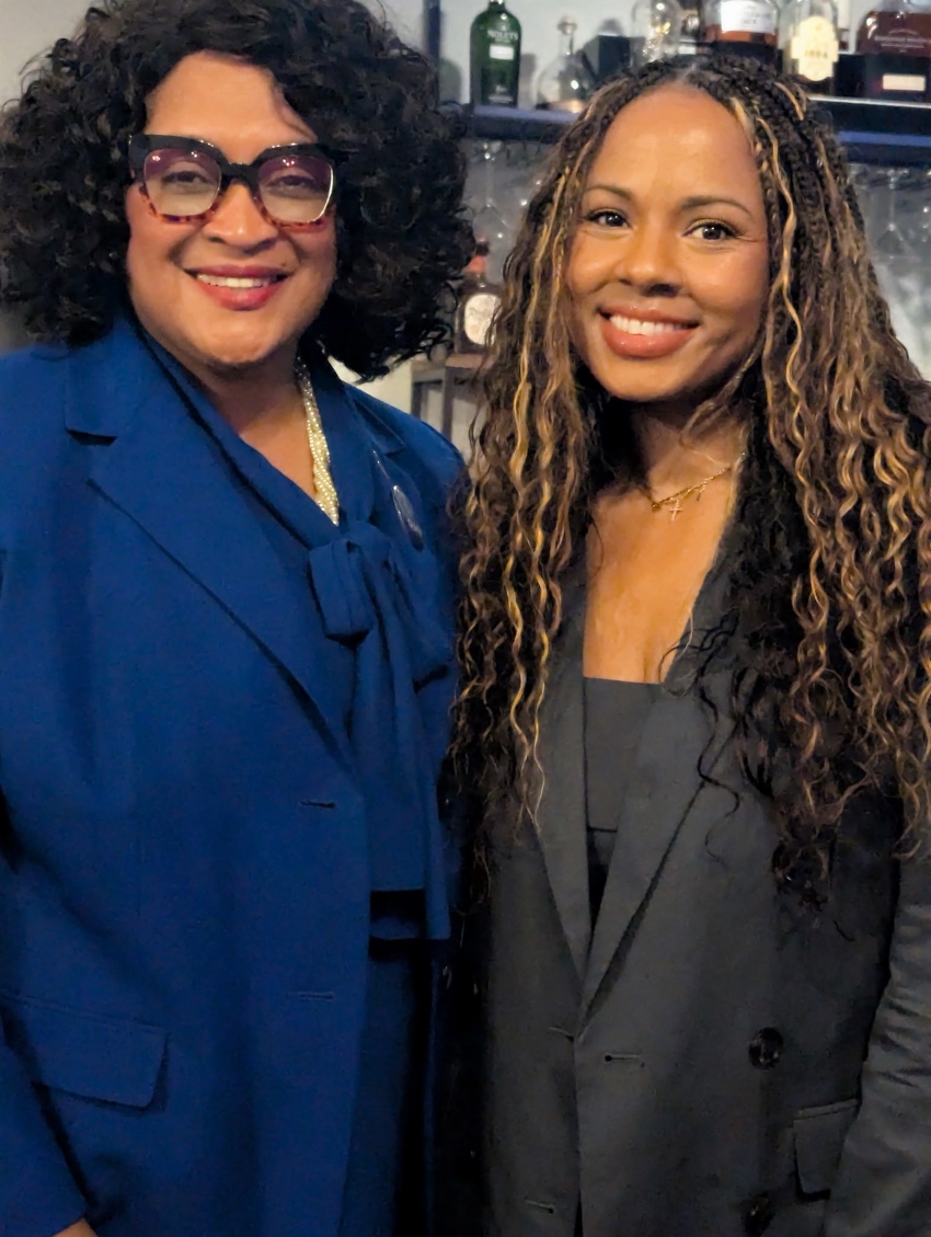 Two women smiling, one with curly hair and glasses wearing a blue blazer, the other with long wavy hair in a dark blazer.