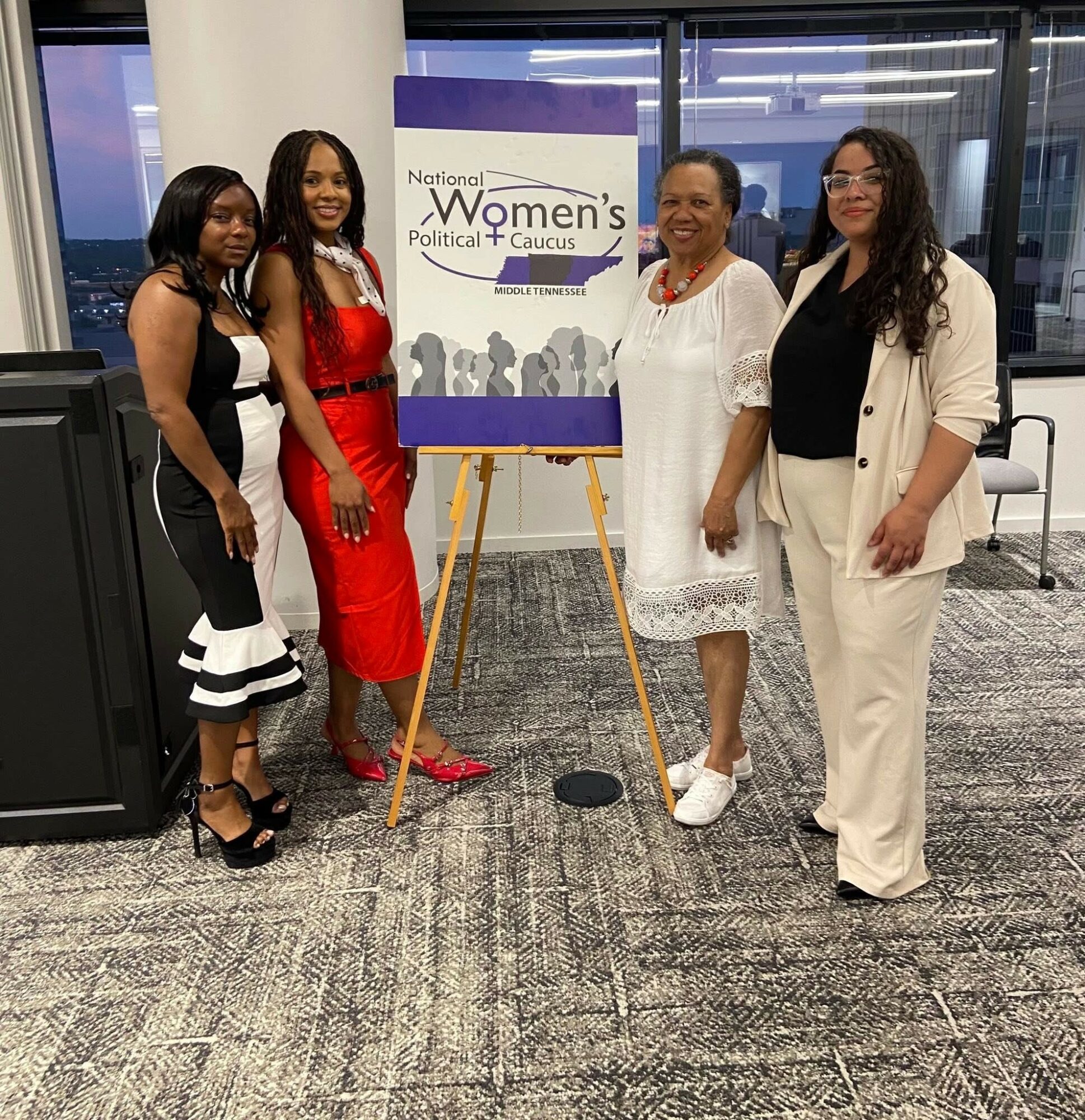 Four women stand together indoors near a sign for Women's Political Caucus, smiling, with large windows behind them.