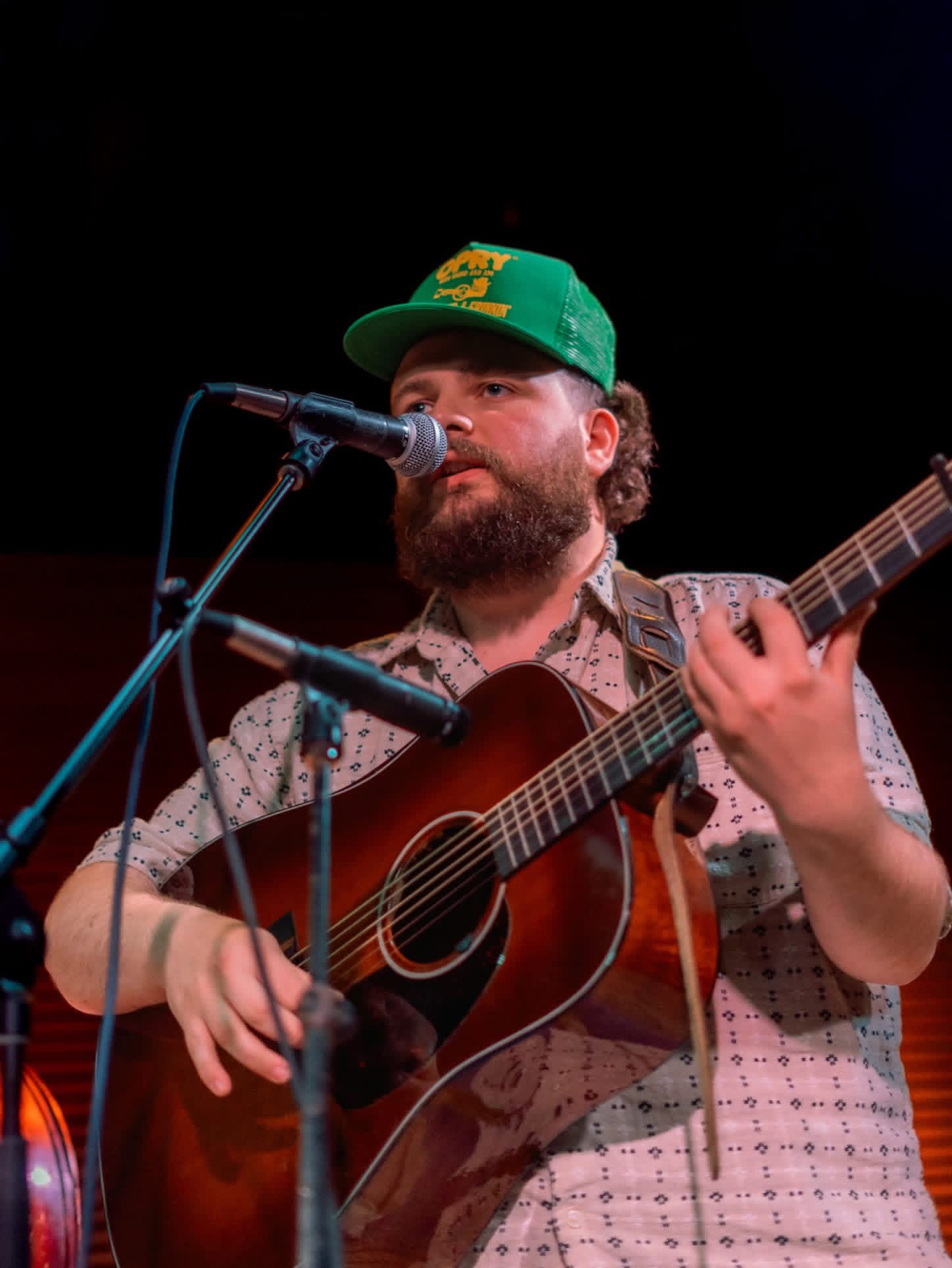 Person with a beard wearing a green cap, playing an acoustic guitar and singing into a microphone.