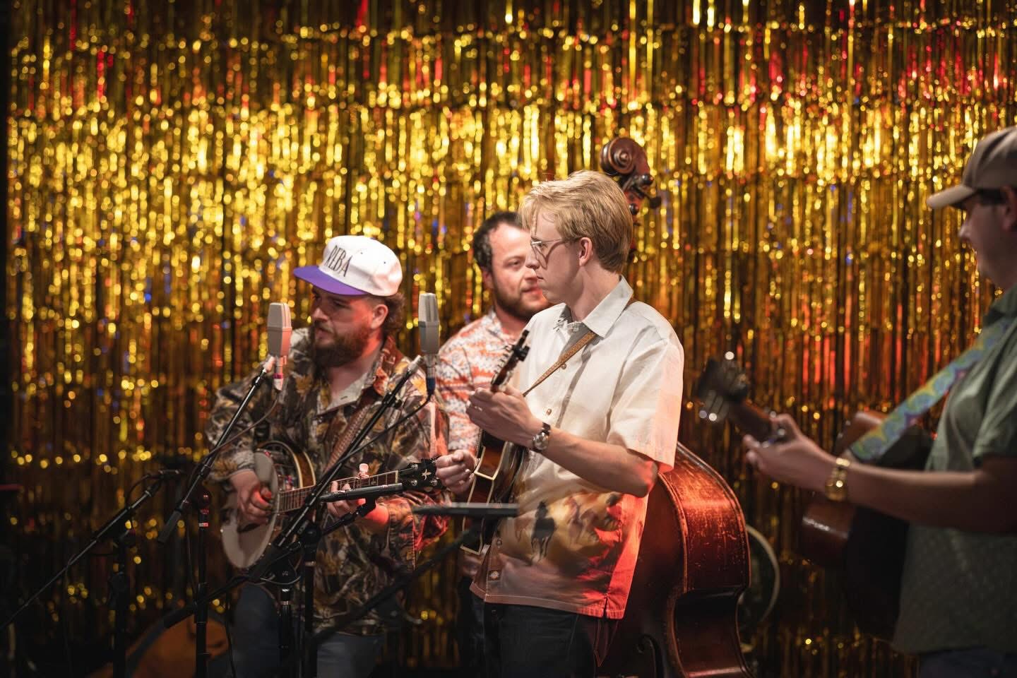 Group of four people performing music on stage with a gold shimmering backdrop.