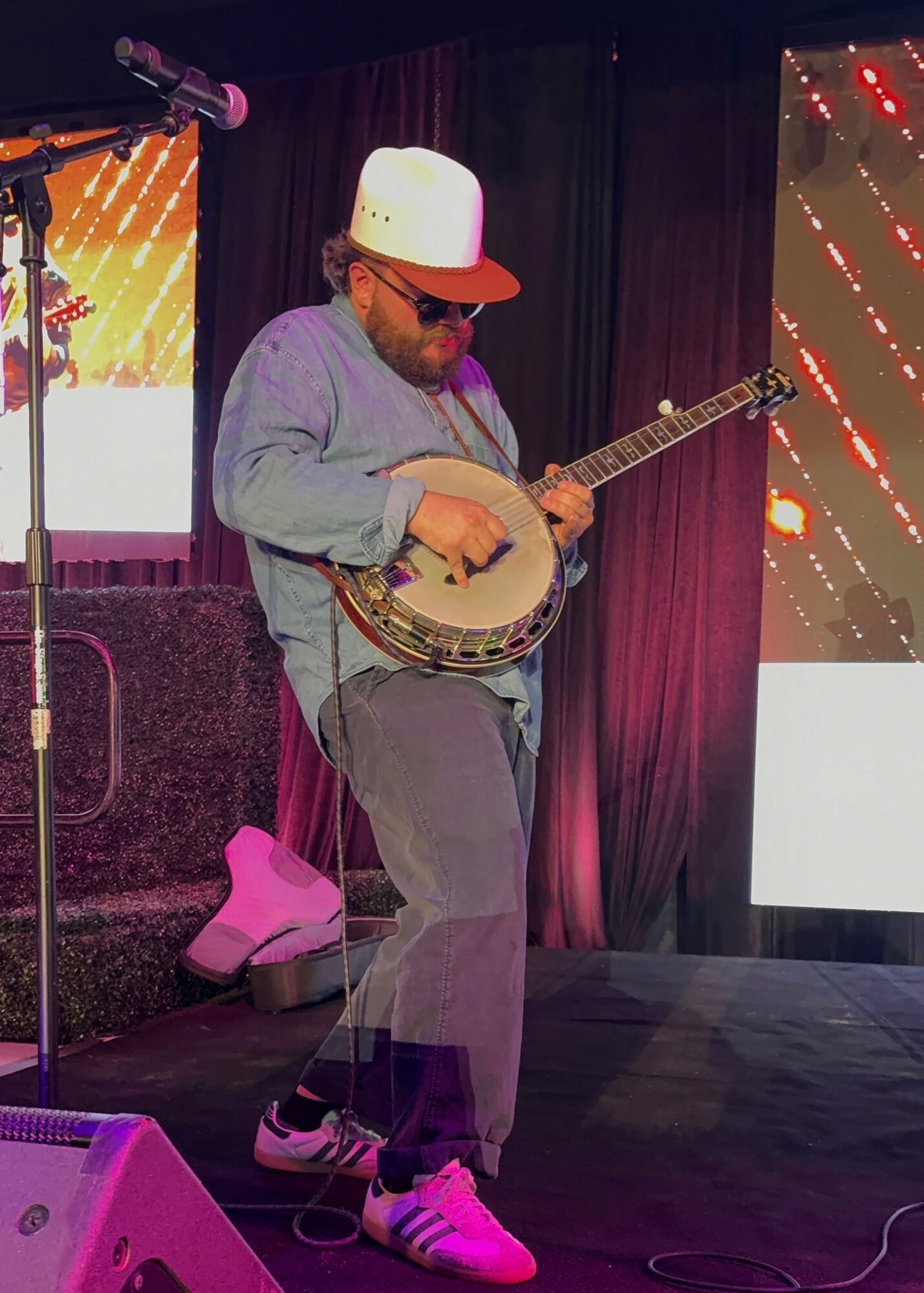 Person playing banjo on stage, wearing a white cap, glasses, gray hoodie, and gray pants, with a microphone nearby.
