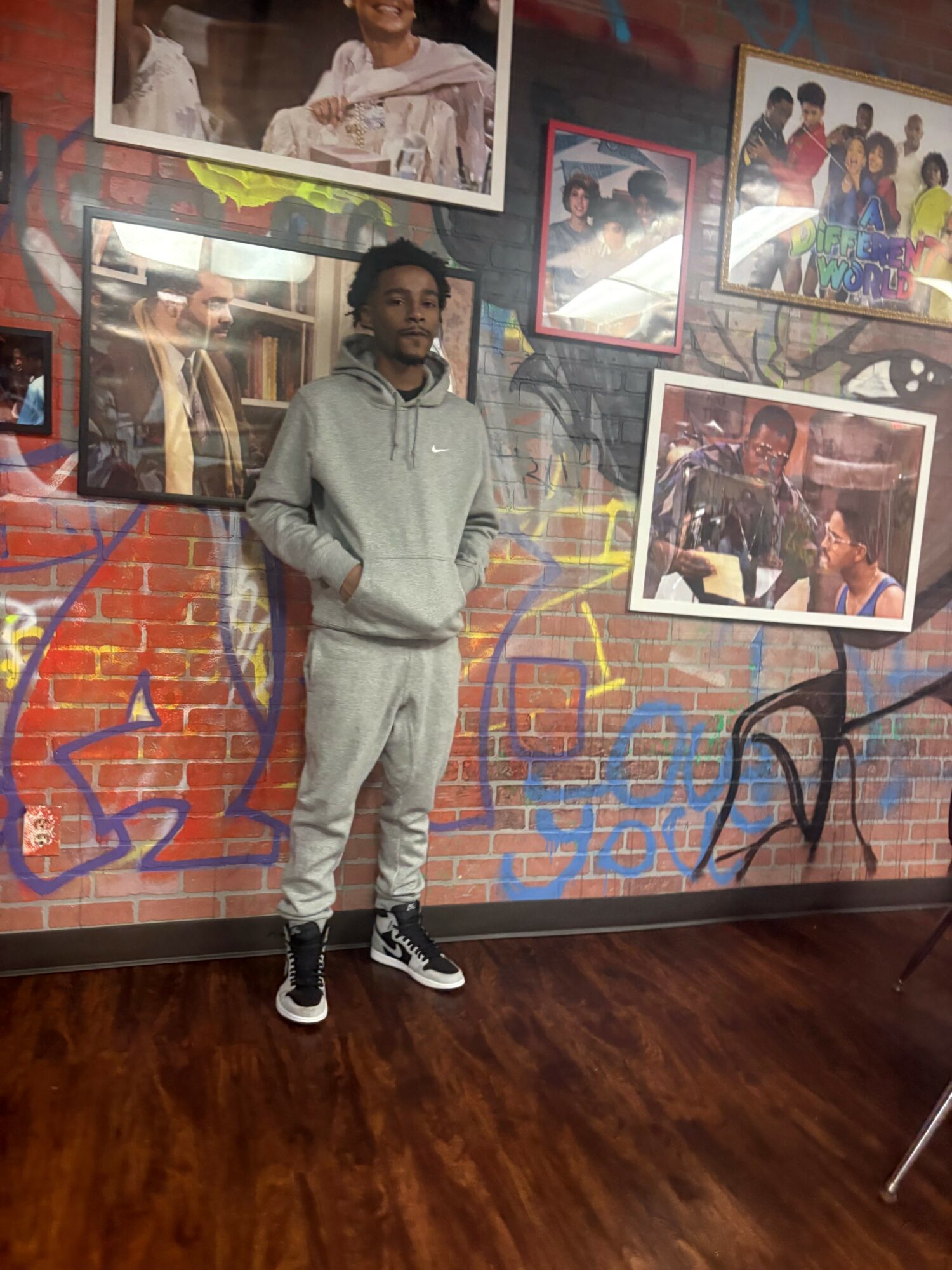 Young man in gray hoodie and sweatpants standing in front of a colorful wall with framed photos, wooden floor beneath him.