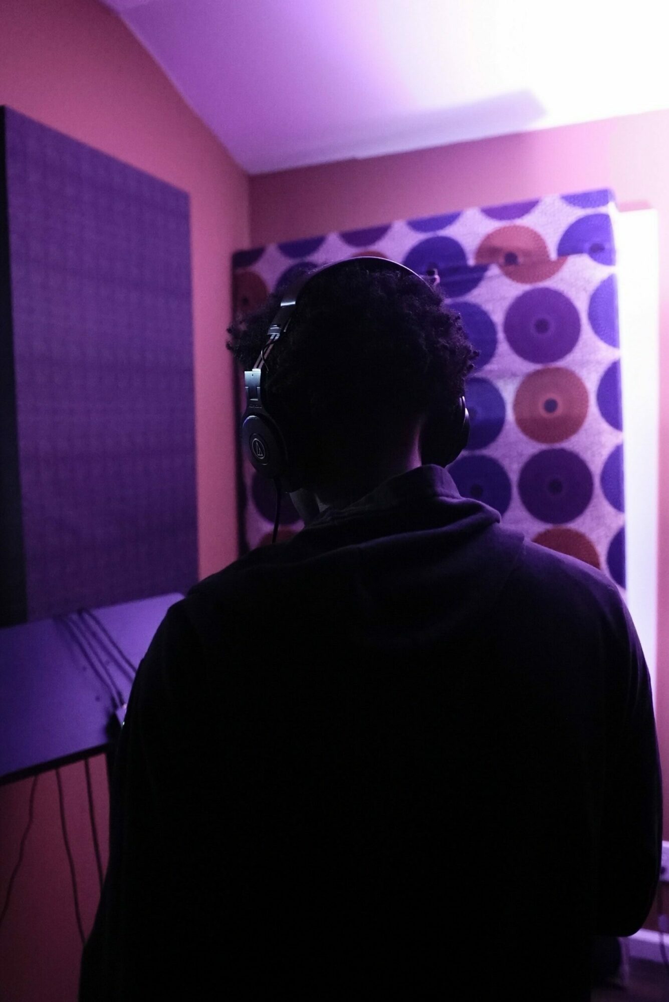 Person with headphones in a room with purple and pink lighting, patterned wall behind them.