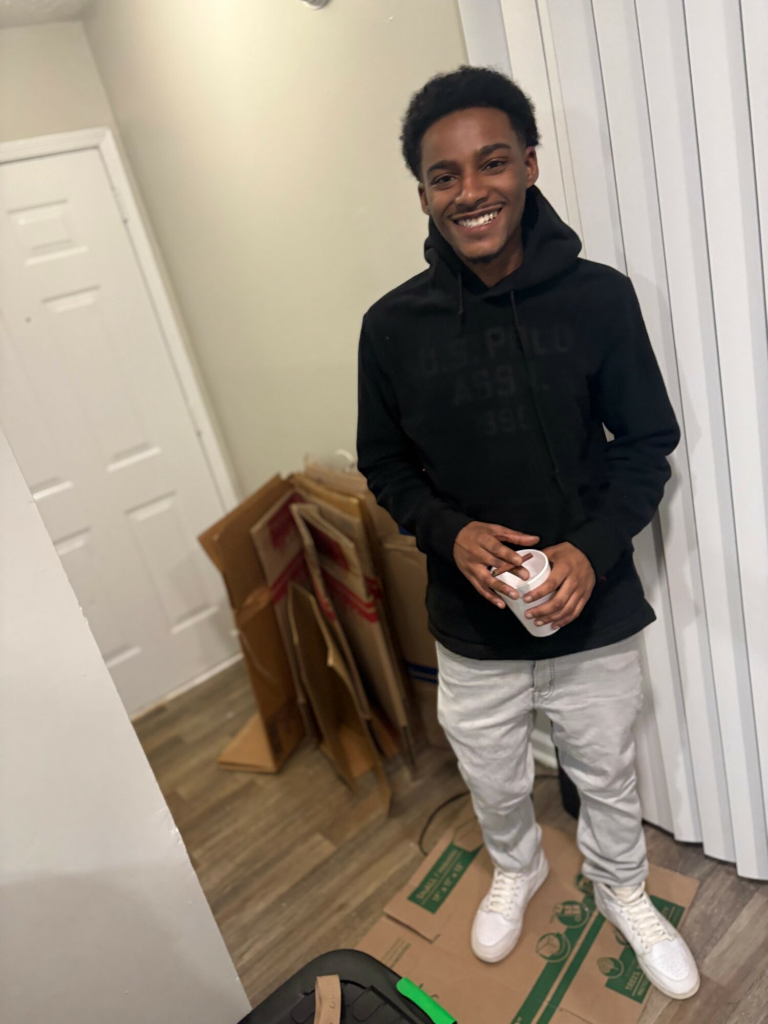 Young person smiling, standing indoors, holding a cup, wearing a black hoodie and light-colored pants, with a door and stacked items in background.