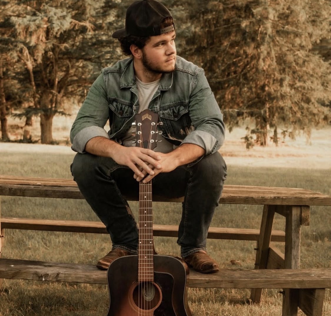 Young man sitting on a wooden bench outdoors with a guitar between his legs, looking to the side, surrounded by trees.