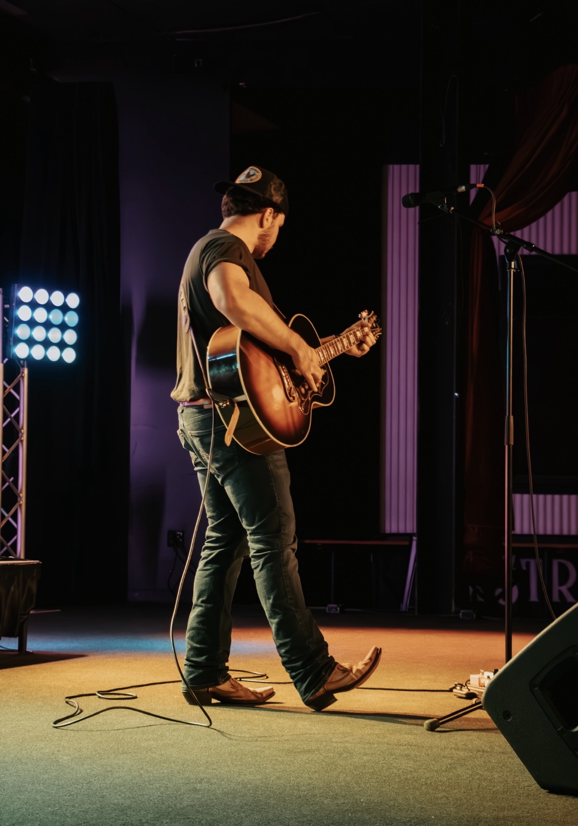 Person playing guitar on stage with stage lights and microphone stand, facing left, wearing a hat and jeans.